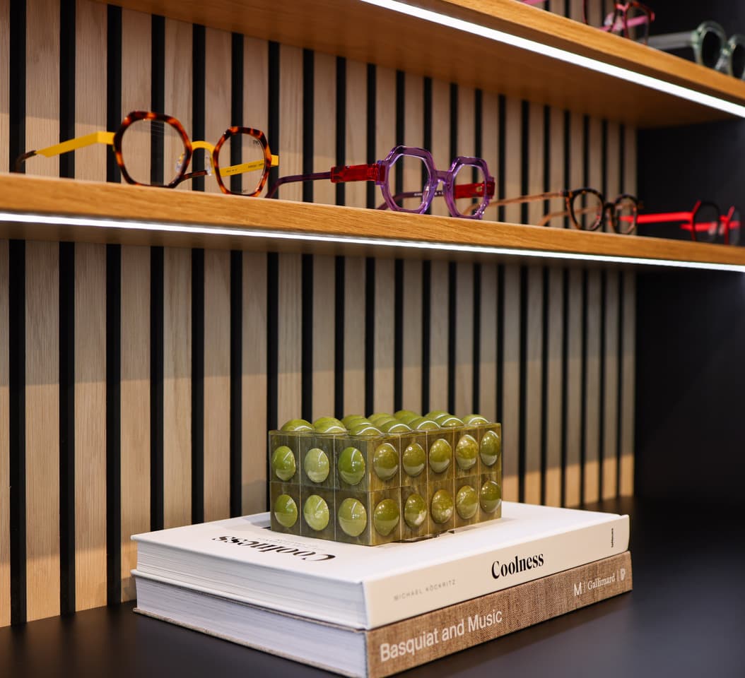Close view of hand-finished acetate frames on an oak shelf in tortoise and amber, violet and red, above a stack of art books.