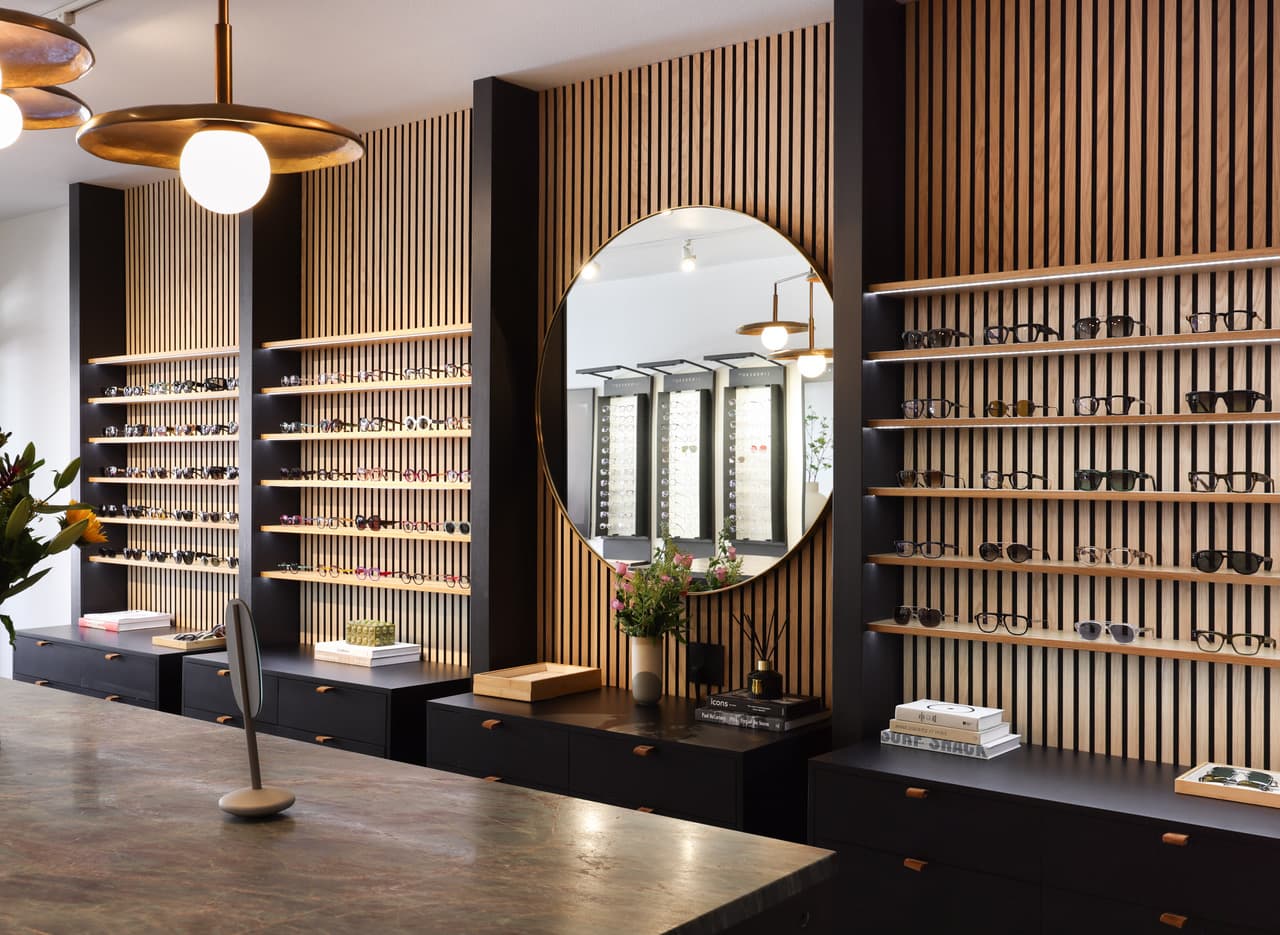 The Eyes On Wall boutique interior: slatted oak walls lined with eyewear, brass pendant lights, a round mirror, and a stone counter.