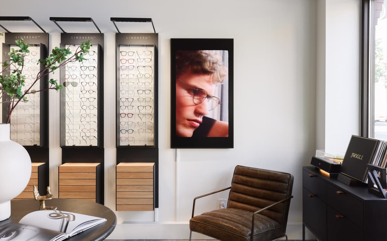 Lit display cases of thin titanium frames beside a framed portrait, a leather lounge chair, and a record player.